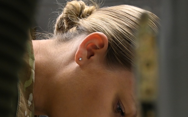 A female military service member wearing MultiCam/OCP camouflage uniform is photographed in close profile view, looking downward with focused concentration. She has blonde hair pulled back in a bun and wears a small stud earring. The image appears to be a candid documentary-style military photograph.