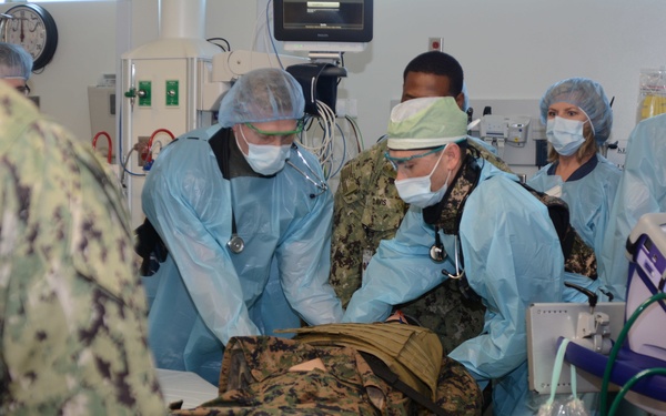 A military medical team in blue surgical gowns, masks, and protective eyewear is treating a patient in military camouflage uniform on a gurney in what appears to be a medical treatment facility or trauma bay. Multiple personnel, including individuals in Navy and Marine Corps uniforms, are actively engaged in patient care. Medical equipment including monitors, IV bags, and various devices are visible throughout the scene.