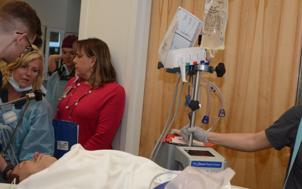 A medical simulation or training exercise is taking place in what appears to be a hospital or clinical training environment. A medical mannequin is lying on a gurney connected to various monitoring devices and IV lines, while several healthcare professionals and students observe and interact with the equipment. A rapid infuser device is visible, and participants wearing scrubs, lanyards, and ID badges suggest this is an educational or demonstration setting.