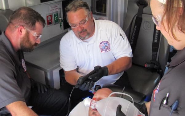 Three emergency medical personnel are treating a patient inside an ambulance. Two responders wear gray uniforms while one wears a white shirt with an EMS patch. The patient is supine and receiving airway management, including what appears to be bag-valve-mask (BVM) ventilation.