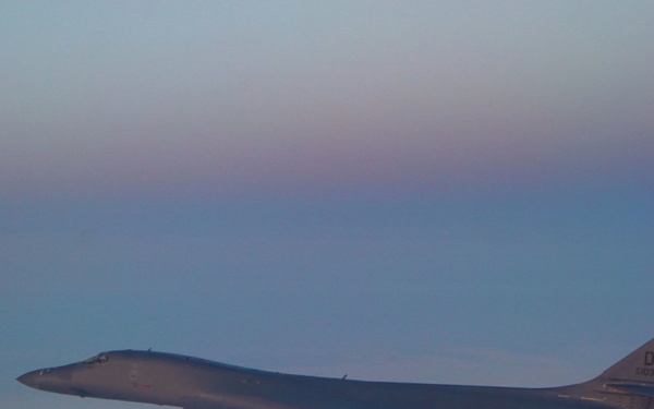 A U.S. Air Force B-1B Lancer strategic bomber is photographed in flight during twilight or dawn conditions. The aircraft bears the tail code 'DY', indicating assignment to Dyess Air Force Base, Texas. It is flying at altitude over what appears to be a snow-covered or icy landscape below.