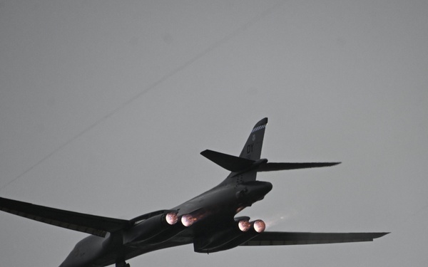 A dark-colored military bomber aircraft is photographed in flight against an overcast gray sky. The aircraft displays visible engine afterburner glow from its four engines, suggesting high-thrust operation. The variable-sweep wing configuration and overall silhouette are consistent with a B-1 Lancer strategic bomber.