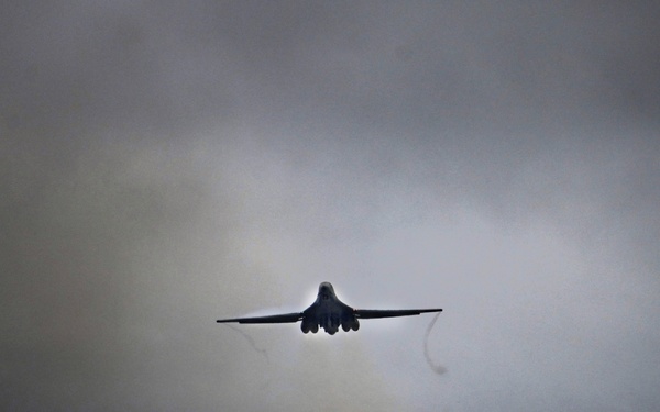 A military aircraft, consistent with the silhouette of a B-1B Lancer strategic bomber, is captured during takeoff or low-altitude flight over a runway in overcast, stormy conditions. The aircraft is viewed from a rear-facing angle, with its variable-sweep wings visible in a swept-back configuration. Runway lighting in green, red, and white is visible below against a backdrop of dark treeline and heavy cloud cover.