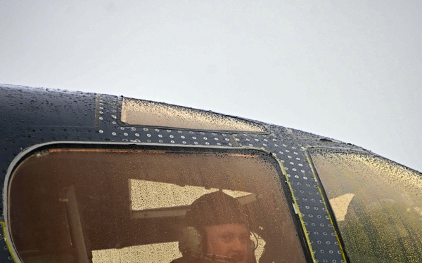 A smiling pilot or crew member is visible through the side window of a large, dark-painted military aircraft. The aircraft fuselage shows riveted metal construction with a rain-covered surface, suggesting wet weather conditions. A second crew member is partially visible through the adjacent canopy section.