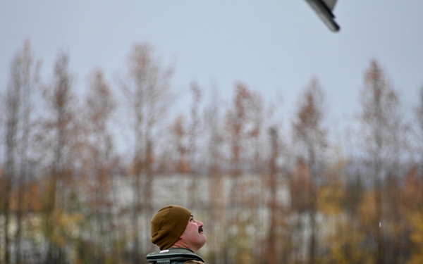 A military aviator or crew member stands on a flight line tarmac, looking upward toward an aircraft wing visible in the upper portion of the frame. The individual is wearing a flight suit, tactical vest, and brown knit cap, suggesting cold-weather operations. The setting appears to be an airfield during autumn, with bare and partially-leafed trees visible in the background under an overcast sky.