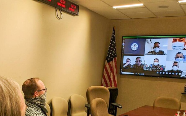 A video teleconference meeting is taking place in what appears to be a U.S. military or government conference room. Two civilians in the foreground are participating in a virtual meeting with multiple military personnel visible on large wall-mounted screens and a laptop. Several participants, both in-person and remote, are wearing face masks, suggesting this occurred during the COVID-19 pandemic period.