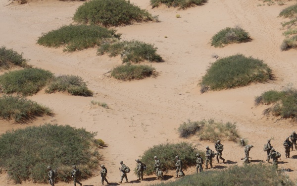 An aerial photograph shows a column of military personnel moving in a single-file patrol formation across a sandy desert terrain. The soldiers are wearing camouflage uniforms and carrying full combat gear and backpacks. The landscape is characterized by sandy soil interspersed with low desert scrub vegetation.