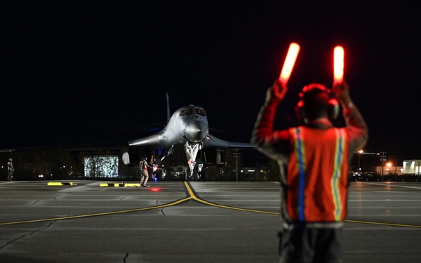 A ground crew member is marshalling a military jet aircraft on a tarmac at night using illuminated red signal wands. The scene takes place on a well-marked airfield apron with yellow taxiway lines visible on the dark pavement. Additional personnel are visible in the background near the aircraft.
