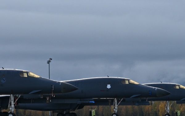 Three dark gray military bomber aircraft are parked on a wet tarmac apron, aligned in a row facing the same direction. The aircraft appear to be B-1B Lancer supersonic bombers, identifiable by their distinctive swept-wing design and elongated fuselages. The setting appears to be a northern or Alaskan airbase, suggested by the autumn foliage and overcast, stormy sky in the background.