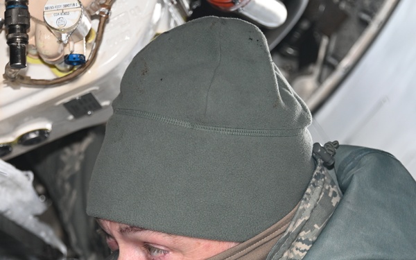 A military technician wearing cold-weather gear, including a fleece balaclava, beanie cap, tactical gloves, and ABU (Airman Battle Uniform) camouflage, is performing maintenance work on what appears to be a military aircraft. The individual is closely inspecting or working on internal mechanical components within an access panel or bay of the aircraft. The cold-weather clothing suggests operations in a low-temperature environment.