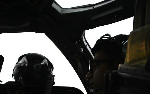 The image shows two aircrew members seated in a military aircraft cockpit, silhouetted against bright exterior light coming through the windscreen. Both individuals are wearing flight helmets and oxygen masks, consistent with military aviation equipment. The cockpit interior is heavily shadowed, creating a high-contrast silhouette composition.