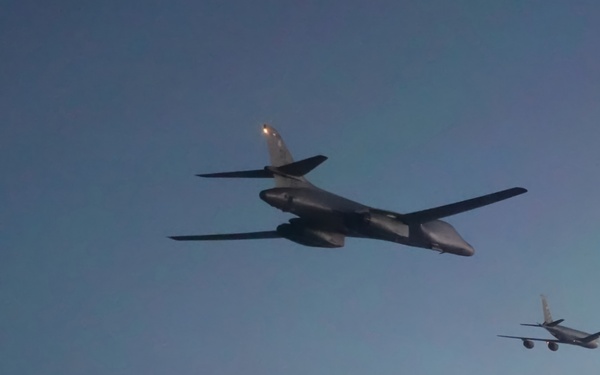 Three military aircraft are flying in a loose formation against a clear twilight sky. The lead aircraft in the foreground appears to be a B-1B Lancer strategic bomber, while the two trailing aircraft in the background appear to be a B-52 Stratofortress and what may be a tanker or additional bomber. The lighting suggests either dawn or dusk conditions, with navigation/anti-collision lights visible on the aircraft.