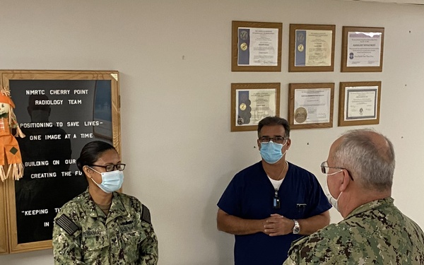 The image shows a group of individuals in a military medical facility hallway, identified by the bulletin board as 'NMRTC Cherry Point Radiology Team.' Two personnel are wearing U.S. Navy digital camouflage uniforms (NWUs) and one individual is wearing navy blue medical scrubs, all wearing blue surgical face masks. A briefing or informal discussion appears to be taking place.