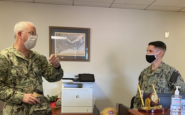 Two U.S. Navy personnel in NWU Type III (digital camouflage) uniforms are engaged in conversation inside a military office setting. Both individuals are wearing face masks, consistent with COVID-19 precautionary protocols. One person appears to be standing and speaking while the other is seated behind a reception or administrative desk.