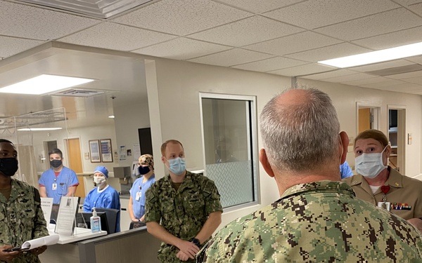A group of U.S. Navy personnel in NWU (Navy Working Uniform) camouflage are gathered in what appears to be a military medical facility or hospital ward. Several civilian medical staff in scrubs are visible in the background near a nurses' station. All individuals are wearing face masks, suggesting this was taken during the COVID-19 pandemic period.