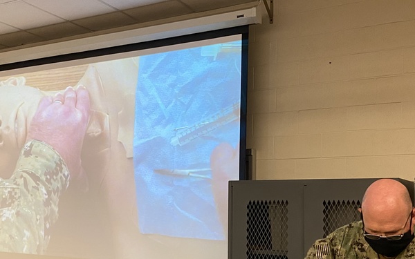A U.S. Navy servicemember wearing a face mask and NWU Type III camouflage uniform is conducting a medical training demonstration in a classroom setting. The instructor is working with a medical training mannequin/simulator while a projected screen behind him displays a close-up instructional video of the same or similar procedure. The setting appears to be a combat casualty care or tactical medicine training course.