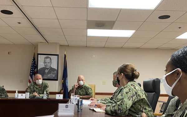 A military briefing or meeting is taking place in a formal conference room with multiple uniformed personnel seated around a large wooden table. All attendees are wearing face masks, suggesting this occurred during the COVID-19 pandemic period. Personnel appear to be from the U.S. Navy and possibly other branches, with documents and name placards visible on the table.