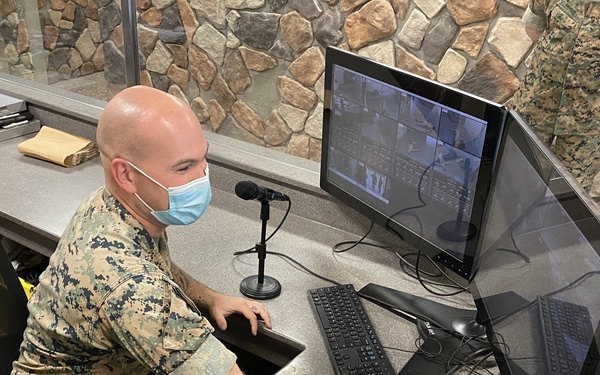 A U.S. Marine in digital camouflage (MARPAT) uniform is seated at a security or operations desk, wearing a blue surgical face mask. He is operating a dual-monitor workstation displaying what appears to be a multi-camera surveillance feed. The setting appears to be a military installation checkpoint or security operations center.