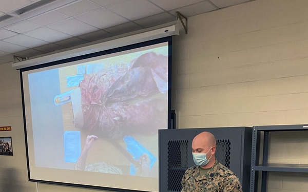 A U.S. Navy corpsman wearing a surgical mask and camouflage uniform conducts a combat casualty care (TCCC) training session in a classroom setting. He is demonstrating medical procedures using a realistic trauma training manikin/simulator on a wooden table, with a projected medical image visible on the screen behind him. Additional training equipment including a CPR manikin head is visible on the floor nearby.