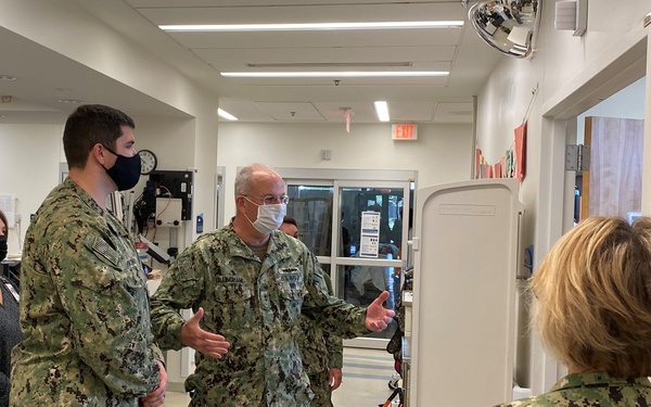 Several U.S. Navy personnel in NWU Type I (digital camouflage) uniforms are gathered in what appears to be a military medical facility or clinic hallway. A senior officer appears to be conducting a briefing or tour, gesturing while speaking to colleagues. All military personnel and at least one civilian are wearing face masks, suggesting this was taken during the COVID-19 pandemic period.