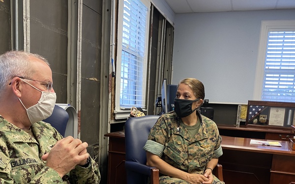 Two military personnel in camouflage uniforms are seated in blue leather chairs in what appears to be a military office, engaged in conversation. Both individuals are wearing face masks. The person on the left has a name tape reading 'GILLINGHAM' and is wearing Navy Working Uniform (NWU), while the person on the right appears to be wearing Marine Corps Combat Utility Uniform (MCCUU).