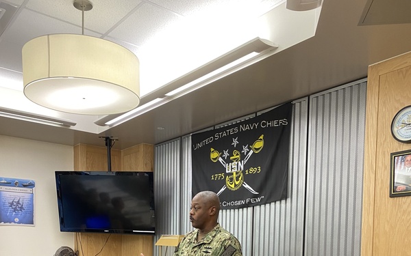 A U.S. Navy service member in NWU Type III (digital camouflage) uniform is speaking or presenting in what appears to be a Chief Petty Officers' Mess or lounge area. Behind the speaker hangs a black 'United States Navy Chiefs' banner with the USN anchor insignia and the dates 1775 and 1893, along with the motto 'The Chosen Few.' The room is furnished with wooden furniture, a wall-mounted television, and various Navy-related decorations.