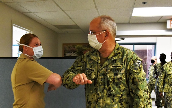 A U.S. Navy officer in NWU Type III camouflage uniform performs an elbow bump greeting with a female service member in khaki uniform, both wearing face masks, in what appears to be a military medical or administrative facility hallway. Additional uniformed personnel are visible in the background walking through the corridor. The scene reflects COVID-19 era health protocols within a military setting.