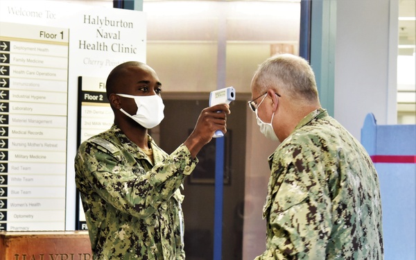 Two U.S. Navy personnel in NWU Type III (digital camouflage) uniforms are shown inside the Halyburton Naval Health Clinic at Cherry Point. One service member is using a non-contact infrared thermometer to screen the other individual's forehead temperature. Both individuals are wearing white face masks, consistent with COVID-19 health screening protocols.