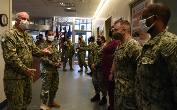 A group of U.S. Navy personnel in digital camouflage uniforms are gathered in what appears to be a military medical facility hallway. A senior officer on the left appears to be addressing or briefing the assembled personnel, all of whom are wearing face masks. The setting suggests a command visit or briefing during the COVID-19 pandemic era.