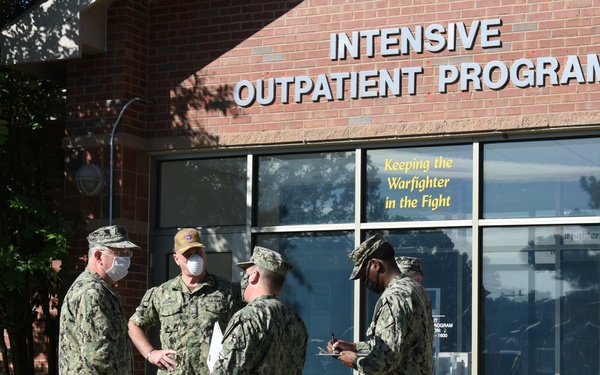 Four U.S. Navy personnel in NWU Type III (woodland camouflage) uniforms are gathered outside a brick building marked 'Intensive Outpatient Program' with the motto 'Keeping the Warfighter in the Fight' visible on the window. Three of the four individuals are wearing face masks, consistent with COVID-19 era protocols. One service member appears to be taking notes on a clipboard while the others converse.