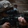 A soldier in digital camouflage uniform and helmet is aiming down the scope of an assault rifle in what appears to be an urban or structured environment. The foreground subject is in sharp focus while additional personnel are visible in the blurred background. The equipment and uniform markings are consistent with U.S. Marine Corps gear.