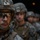 Three military personnel in combat gear are standing in a line, with the foreground soldier in sharp focus and the others progressively blurred in the background. The soldiers are wearing digital camouflage uniforms consistent with U.S. Marine Corps MARPAT pattern, combat helmets with mounted accessories, and tactical vests. The image appears to capture a formation or pre-mission staging moment.