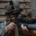 A military service member in MARPAT (Marine pattern) digital camouflage uniform and helmet is aiming a rifle with an underbarrel grenade launcher attachment. The soldier is equipped with multiple optical devices mounted on both the helmet and the weapon. The image appears to be a close-up tactical or training photograph taken in a structured environment.