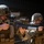 Two military personnel in MARPAT (Marine digital camouflage) uniforms are shown in a firing stance, aiming rifles forward. The foreground soldier is equipped with an M16/M4-style rifle fitted with an M203 grenade launcher, optic scope, and tactical flashlight. The image appears to depict a training exercise or tactical drill.