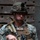 A U.S. Marine in full combat gear is photographed in a portrait-style image against a wooden background. The Marine is wearing MARPAT digital camouflage uniform and helmet, carrying an M4-style carbine with optics and accessories. Name tape and unit patch are visible on the plate carrier.