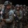 A column of U.S. Marines in full combat gear moves in formation, likely disembarking from a naval vessel's well deck or cargo bay. The lead Marine carries an M4 carbine with accessories and wears MARPAT digital camouflage with a tactical vest and helmet. Additional Marines and military vehicles are visible in the background.