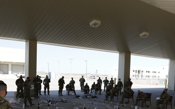 Military personnel in camouflage uniforms are gathered under a large covered outdoor structure, appearing to conduct a tactical planning or briefing exercise. A floor map or terrain model has been laid out on the ground using tape, markers, and labeled placards to represent positions or locations. Some personnel are seated while others stand around the layout, suggesting an operational planning session or rehearsal of concept drill.