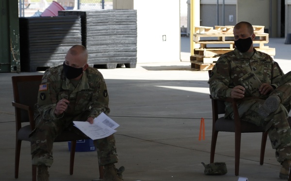 Two U.S. Army soldiers in OCP (Operational Camouflage Pattern) uniforms and black face masks are seated in chairs during what appears to be a military terrain model briefing or operations order (OPORD) exercise. The floor in front of them is marked with a sand table layout featuring numbered placards (41-45), route markers labeled 'MSR FISH' and 'MSR HEN', a 'GATE 2' sign, and colored lines indicating roads or boundaries. One soldier is holding and reviewing printed documents.