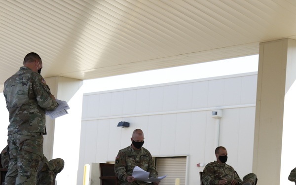 Military personnel in camouflage uniforms and face masks are seated and standing under a covered outdoor area, appearing to participate in a briefing or exercise. Numbered placards and location signs (including 'Camp Bondsteel,' 'Novo Selo,' 'DoG 1,' 'MSR HEN,' and numbers 83-87) are arranged on the ground in front of them. The setting suggests a military planning exercise or map exercise (MAPEX) with markers representing positions or locations.
