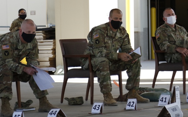Three U.S. Army soldiers in OCP (Operational Camouflage Pattern) uniforms and face masks are observing a large-scale terrain model or sand table exercise on a hangar or warehouse floor. Numbered markers (39-46 visible) and labeled placards including 'MSR FISH' and 'Camp Bondsteel' are arranged on the floor to represent a tactical map layout. A fourth soldier is visible in the background.