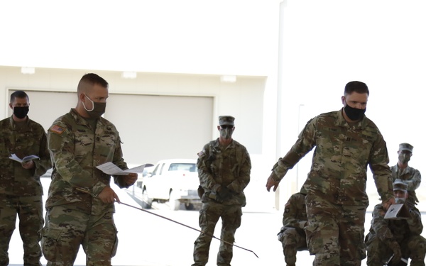 Military personnel in OCP (Operational Camouflage Pattern) uniforms and face masks are conducting what appears to be a terrain model briefing or operational planning exercise on a large paved surface, likely a flight line or hangar apron. Numbered and labeled markers are arranged on the ground to represent a geographic area, with place names such as 'Pristina,' 'Mitrovica,' and 'FHT' visible, suggesting a Kosovo-related operational planning scenario. Several soldiers are walking through the layout while others observe from the sides.