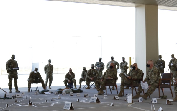 A military briefing or terrain model exercise is taking place in an outdoor covered area, with one soldier using a pointer to brief a group of seated and standing soldiers. A large terrain model or operational map layout is constructed on the ground using numbered markers, labeled tent cards, and drawn lines. The soldiers are wearing camouflage uniforms and face masks, suggesting this occurred during the COVID-19 pandemic period.