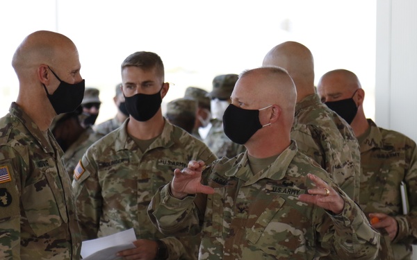 A group of U.S. Army soldiers in Operational Camouflage Pattern (OCP) uniforms are gathered in what appears to be a covered outdoor or hangar-type facility. All personnel are wearing black face masks, consistent with COVID-19 era protocols. A central figure appears to be briefing or conversing with others, using hand gestures to communicate.