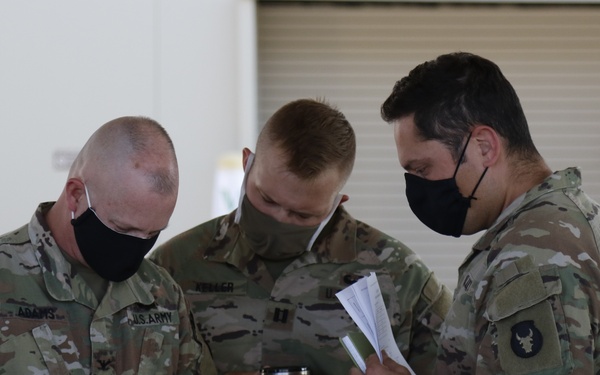 Three U.S. Army soldiers in MultiCam (OCP) uniforms and black face masks are gathered outdoors reviewing documents together. All three wear the same unit patch (appears to be a buffalo/bison insignia) on their left shoulders. The setting appears to be a military installation with garage-style bay doors visible in the background.