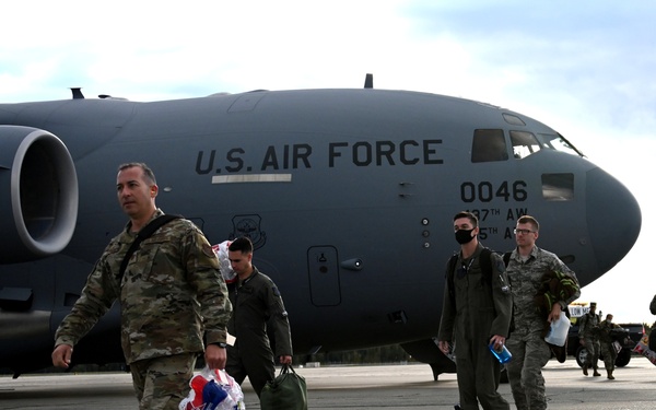Military personnel in camouflage and flight suits are disembarking or walking away from a U.S. Air Force C-17 Globemaster III transport aircraft on a tarmac. The aircraft bears the tail number 0046 and partial unit markings including '37th AW' and '5th AS'. One service member in the foreground is carrying what appears to be patriotic-themed balloons, suggesting a homecoming or welcome event.