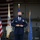 A U.S. Air Force senior non-commissioned officer stands at attention in a military hangar during a formal ceremony. He is wearing the Air Force Service Dress uniform with multiple ribbons and a dark face mask. Three flags are visible behind him: the U.S. flag, the New Jersey state flag, and the U.S. Air Force flag.