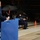 A formal military ceremony is taking place inside an aircraft hangar, with a uniformed Air Force member speaking at a blue podium. In the background, a seated uniformed individual wearing a face mask is applauding, with a military jet fighter visible behind them. The setting and attire suggest an official recognition or retirement ceremony.