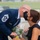A U.S. Air Force senior enlisted member in dress blues, wearing a black face mask, leans in to kiss a woman in a black dress who is holding a bouquet of flowers with a yellow-green ribbon. The rank insignia on his sleeve indicates he holds a Chief Master Sergeant or higher enlisted rank. The setting appears to be an outdoor military ceremony or event, likely a retirement or promotion ceremony.