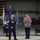 A military ceremony taking place inside an aircraft hangar, likely a retirement or change of command ceremony for a senior non-commissioned officer. Three Air Force personnel in dress blue uniforms are present, with one holding a shadow box display case shaped like a Chief Master Sergeant chevron containing medals, patches, and memorabilia. All visible personnel are wearing face masks, suggesting this occurred during the COVID-19 pandemic period.