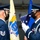 Two U.S. Air Force personnel in dress blues are participating in a formal military ceremony involving a folded American flag. The airman on the right, wearing a 'Base Honor Guard' patch and white gloves, is presenting or transferring a tri-folded U.S. flag to the airman on the left. Both personnel are wearing black face masks, indicating this ceremony took place during the COVID-19 pandemic period.